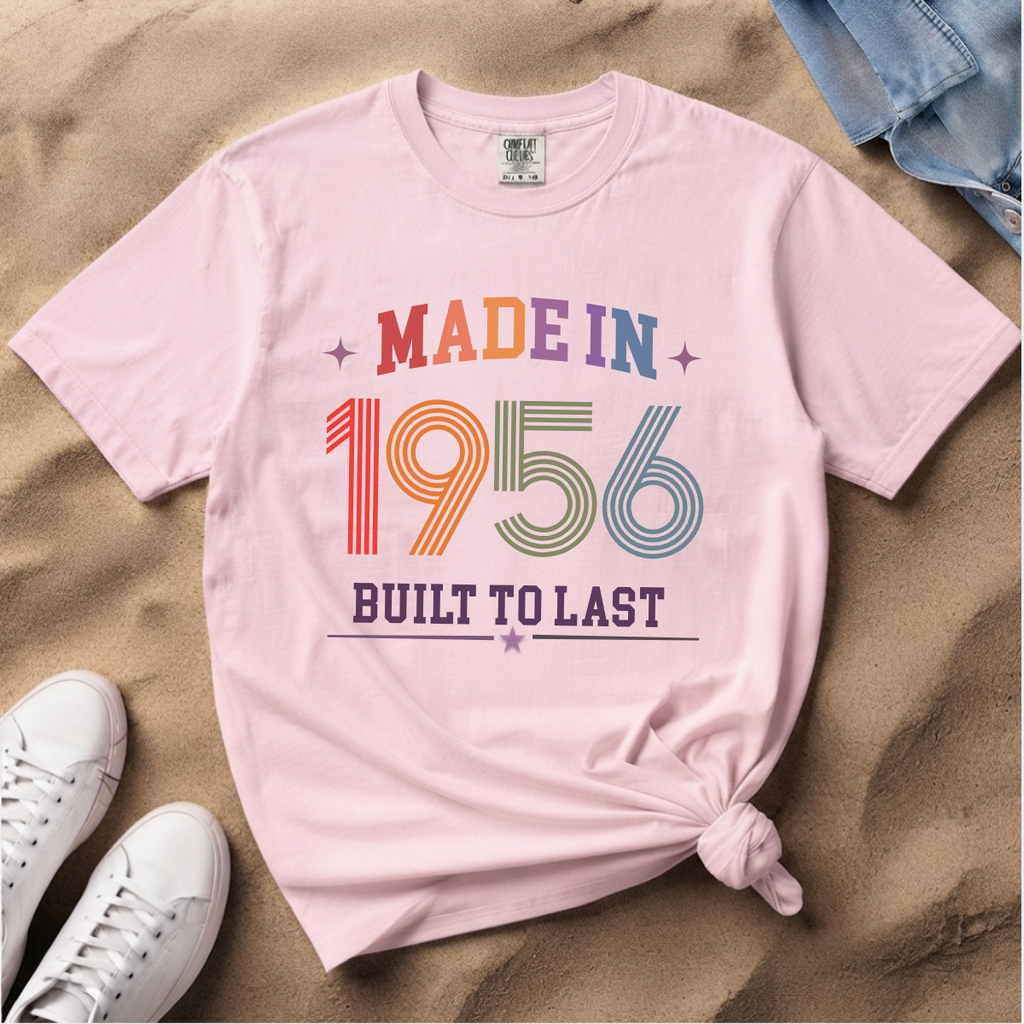 A light pink graphic t-shirt laid flat on a sandy background, styled with white sneakers and a pair of blue jeans. The t-shirt features a retro-style graphic that reads 'MADE IN 1956 BUILT TO LAST' in multicolor lettering.