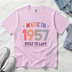 A light pink short-sleeve t-shirt laid flat on a white fabric background, featuring a retro-style multicolor graphic that reads 'MADE IN 1957 BUILT TO LAST' with stars. Styled flat-lay props include a denim jacket corner, white sneakers, and a watch.