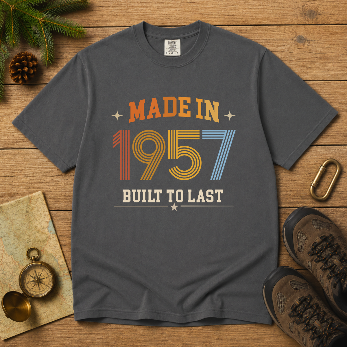 A dark gray graphic t-shirt laid flat on a wooden surface, surrounded by outdoor-themed props including pine branches, a compass, a map, a carabiner, and hiking boots. The shirt features a retro-style graphic that reads 'MADE IN 1957 BUILT TO LAST' in orange, yellow, and blue colors.