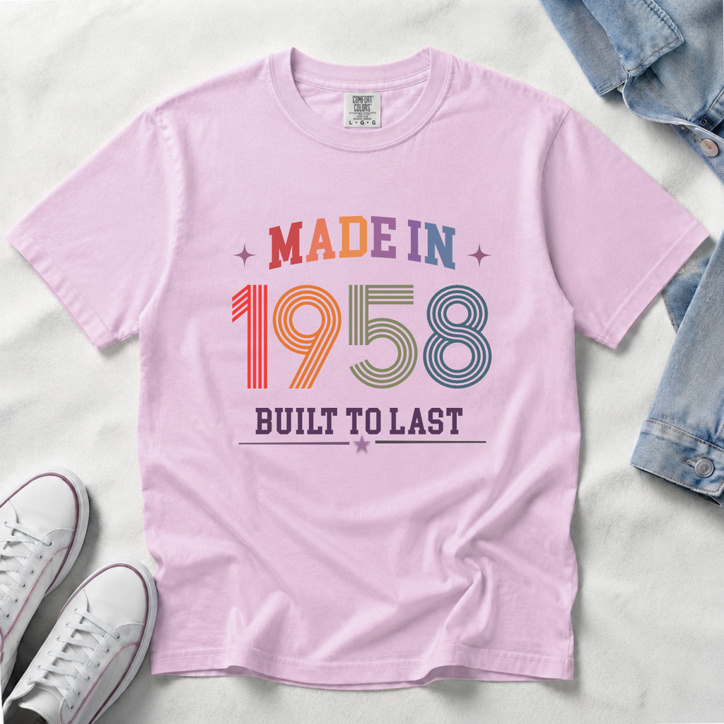 A light pink short-sleeve t-shirt laid flat on a white fabric background, featuring a retro-style multicolor graphic that reads 'MADE IN 1958 BUILT TO LAST' with stars. Styled flat-lay props include folded blue denim at the top right and white sneakers at the bottom left.