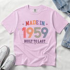 A light pink, orchid short-sleeve t-shirt laid flat on a white fabric background, featuring a retro-style multicolor graphic that reads 'MADE IN 1959 BUILT TO LAST' with stars. Styled flat-lay props include white sneakers at the bottom left, a denim jacket at the top right