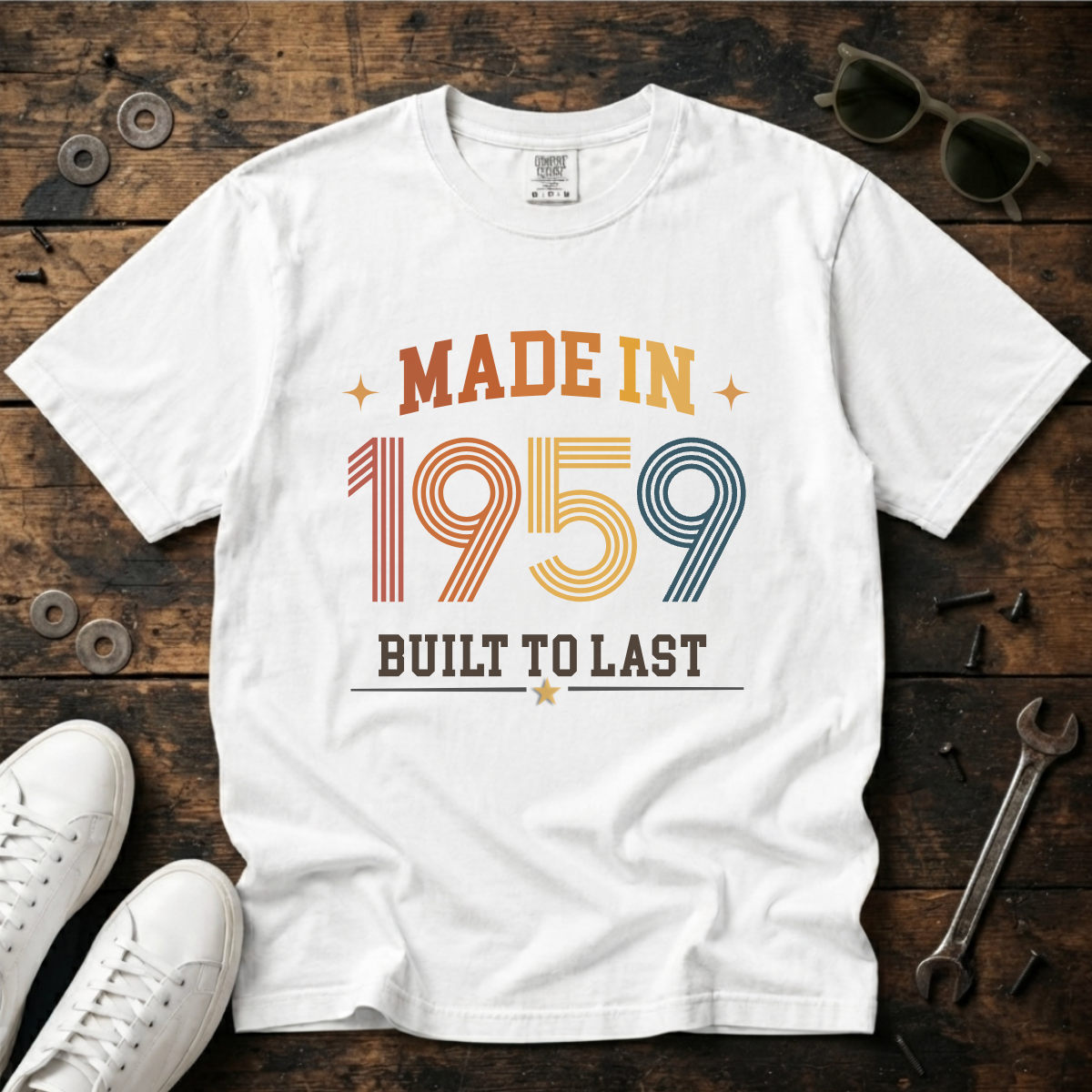 A white short-sleeve graphic T-shirt laid flat on a rustic dark wood surface, surrounded by white sneakers, sunglasses, coins, and a wrench. The shirt reads “MADE IN 1959 BUILT TO LAST” in retro orange, yellow, and blue lettering.