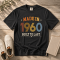 A black graphic t-shirt laid flat on a rustic wooden surface, styled with white sneakers, a notebook, and a sprig of greenery. The t-shirt features a retro-style graphic reading 'MADE IN 1960 BUILT TO LAST' in orange, yellow, blue, and white colors.