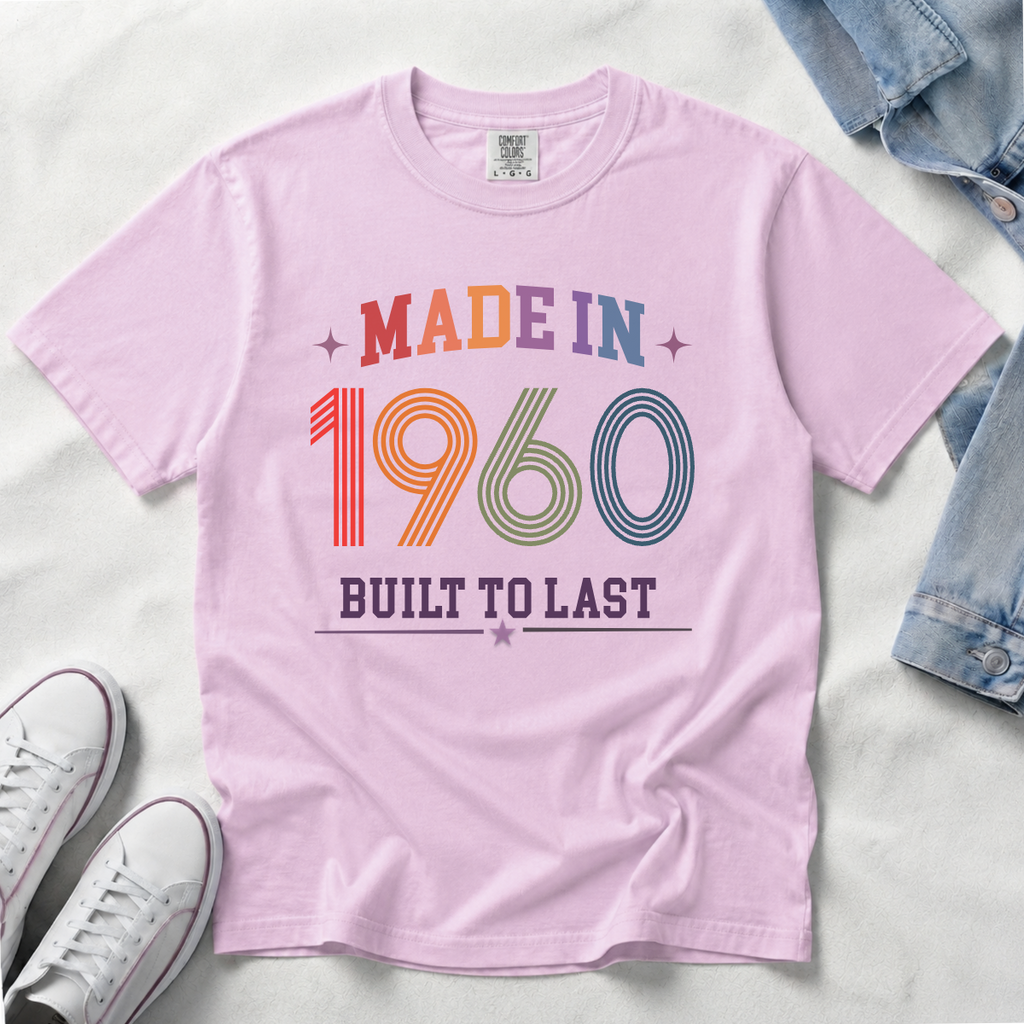 A light pink short-sleeve t-shirt laid flat on a white fabric background, featuring a retro-style multicolor graphic that reads 'MADE IN 1960 BUILT TO LAST' with stars. Styled flat-lay props include folded blue denim at the top right and white sneakers at the bottom left.