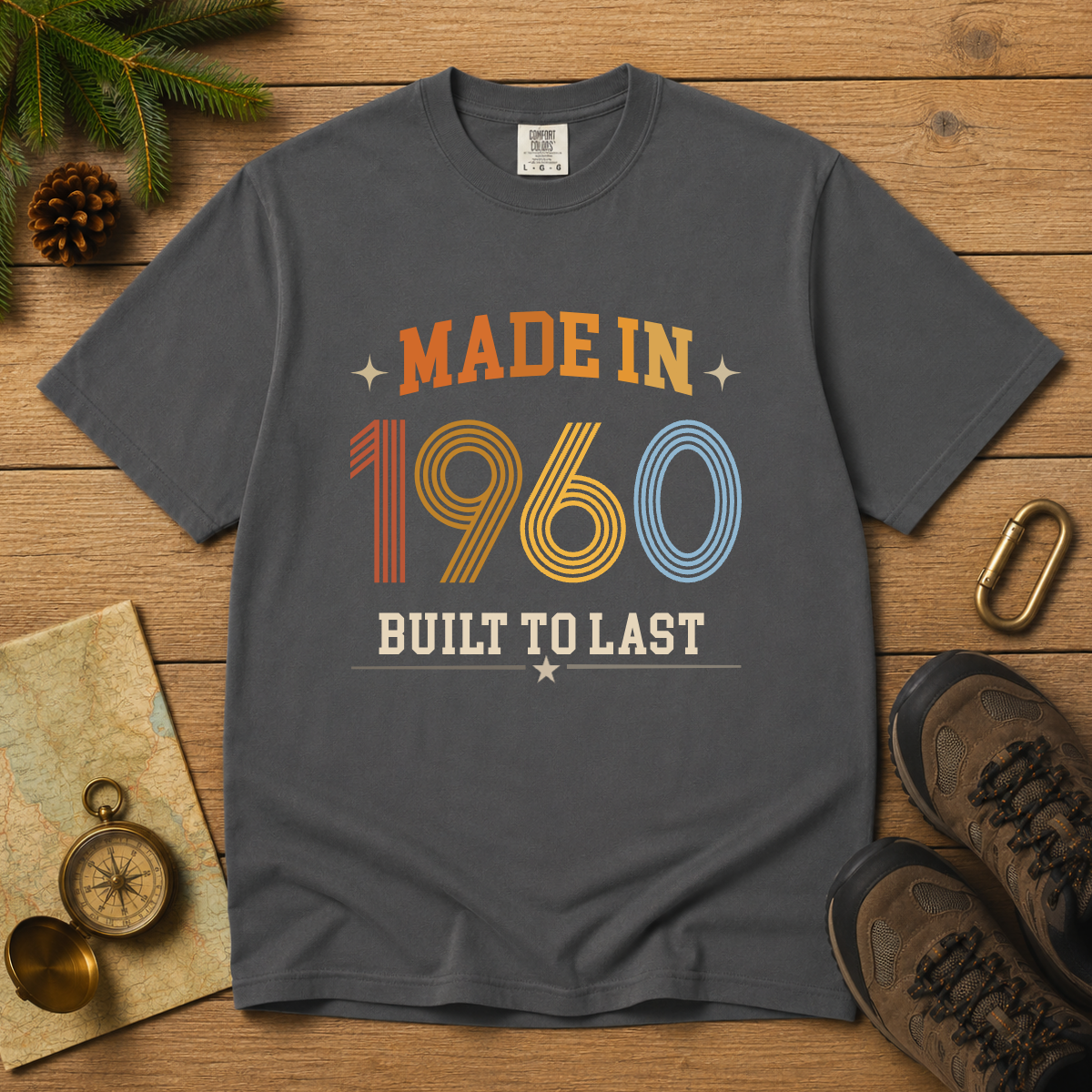 A dark gray graphic t-shirt laid flat on a wooden surface, surrounded by vintage-style props including pine branches, a compass, a map, a carabiner, and hiking boots. The shirt features a retro graphic that reads 'MADE IN 1960 BUILT TO LAST' in orange, yellow, blue, and white.