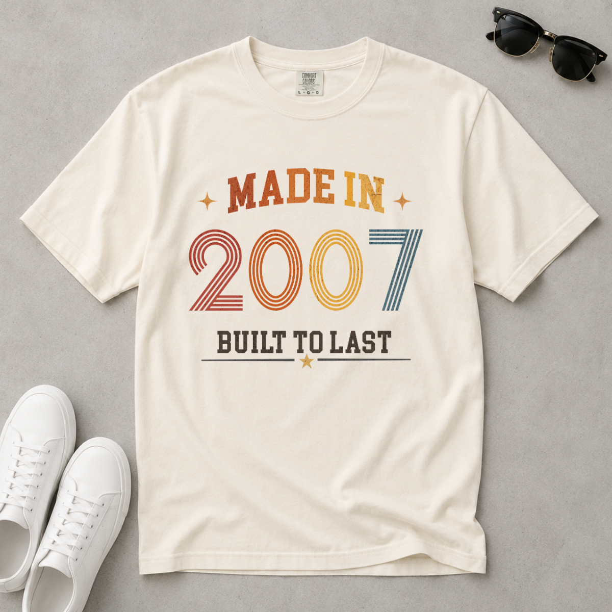 An off-white, ivory graphic t-shirt laid flat on a gray background, styled with white sneakers and black sunglasses. The t-shirt features a retro-style graphic that reads 'MADE IN 2007 BUILT TO LAST' in orange, yellow, and blue colors. The perfect birthday gift for someone born in 2007.
