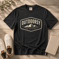 A black graphic t-shirt laid flat on a rustic wooden surface, styled with white sneakers, a notebook, and a sprig of greenery. The t-shirt features a cream-colored retro graphic with mountains and the text 'OUTDOORSY' and 'GO OUTSIDE'.