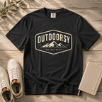 A black graphic t-shirt laid flat on a rustic wooden surface, styled with white sneakers, a notebook, and a sprig of greenery. The t-shirt features a cream-colored retro graphic with mountains and the text 'OUTDOORSY' and 'GO OUTSIDE'.